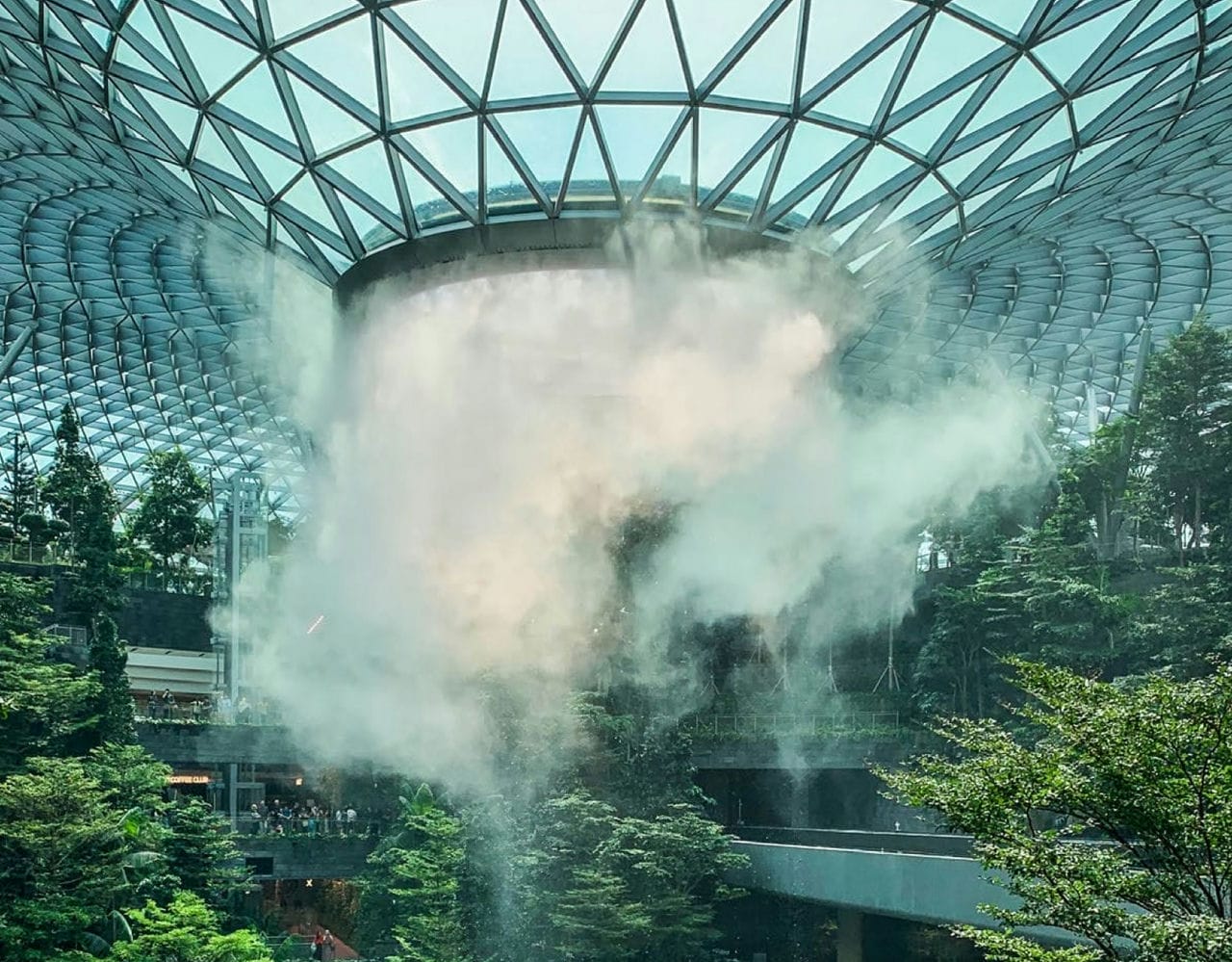 A water fountain below a glass ceiling, surrounded by plants